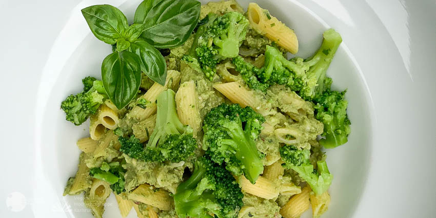 In einem weiß glänzenden Teller liegen die in Sauce gewälzten Nudeln mit Brokkoli und Basilikumblättern als eine in Grün-Tönen schattierte Mahlzeit.