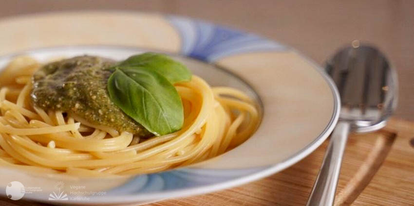 Auf einem Teller sind Spaghetti mit grünem Pesto angerichtet, und mit zwei Basilikumblättern garniert. Der Teller steht auf einem Holzbrettchen, auf dem das Essbesteck liegt. Während die Spaghetti mit dem Pesto im Fokus des Bildes liegen, fällt die Schärfe im Vorder- und Hintergrund sanft ab.